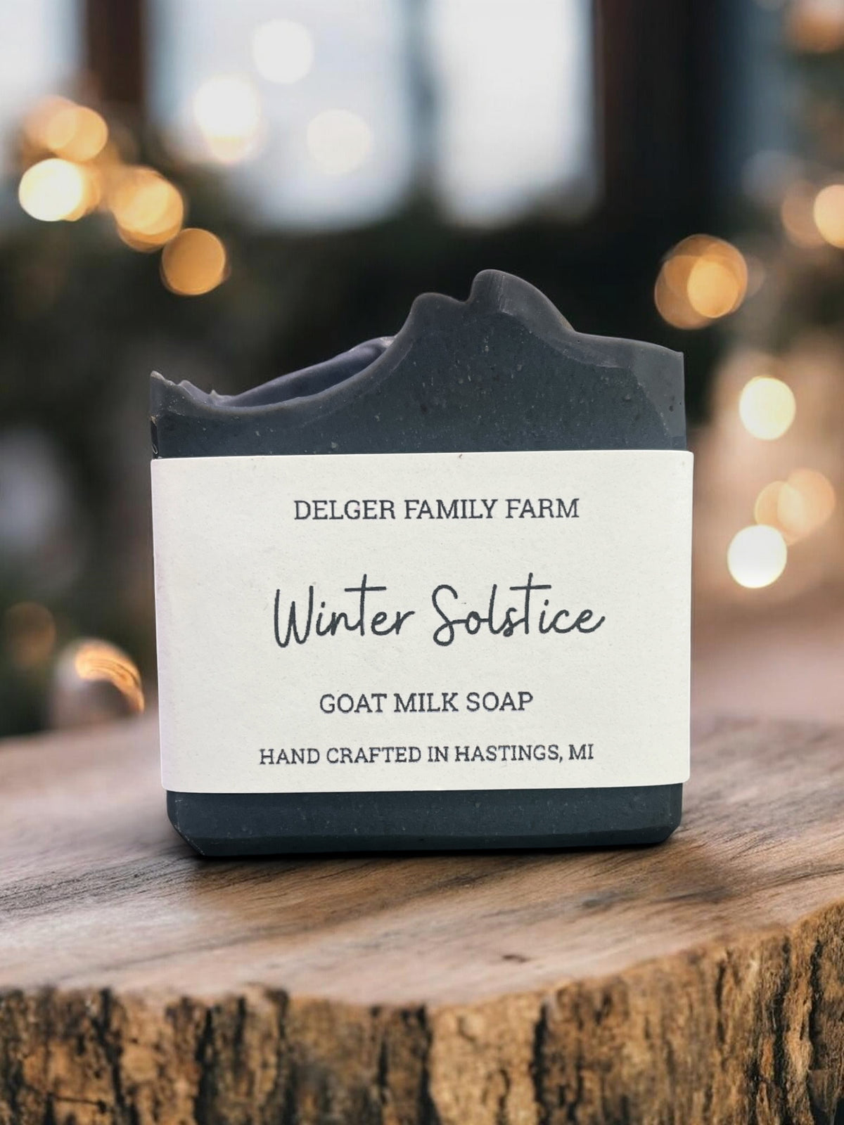 A handmade goat milk soap bar with a rustic, hand-crafted top edge, wrapped in a white paper label that reads 'Winter Solstice' by Delger Family Farm. The soap is displayed on a rustic wooden log slice with a warm, blurred bokeh background of lights.