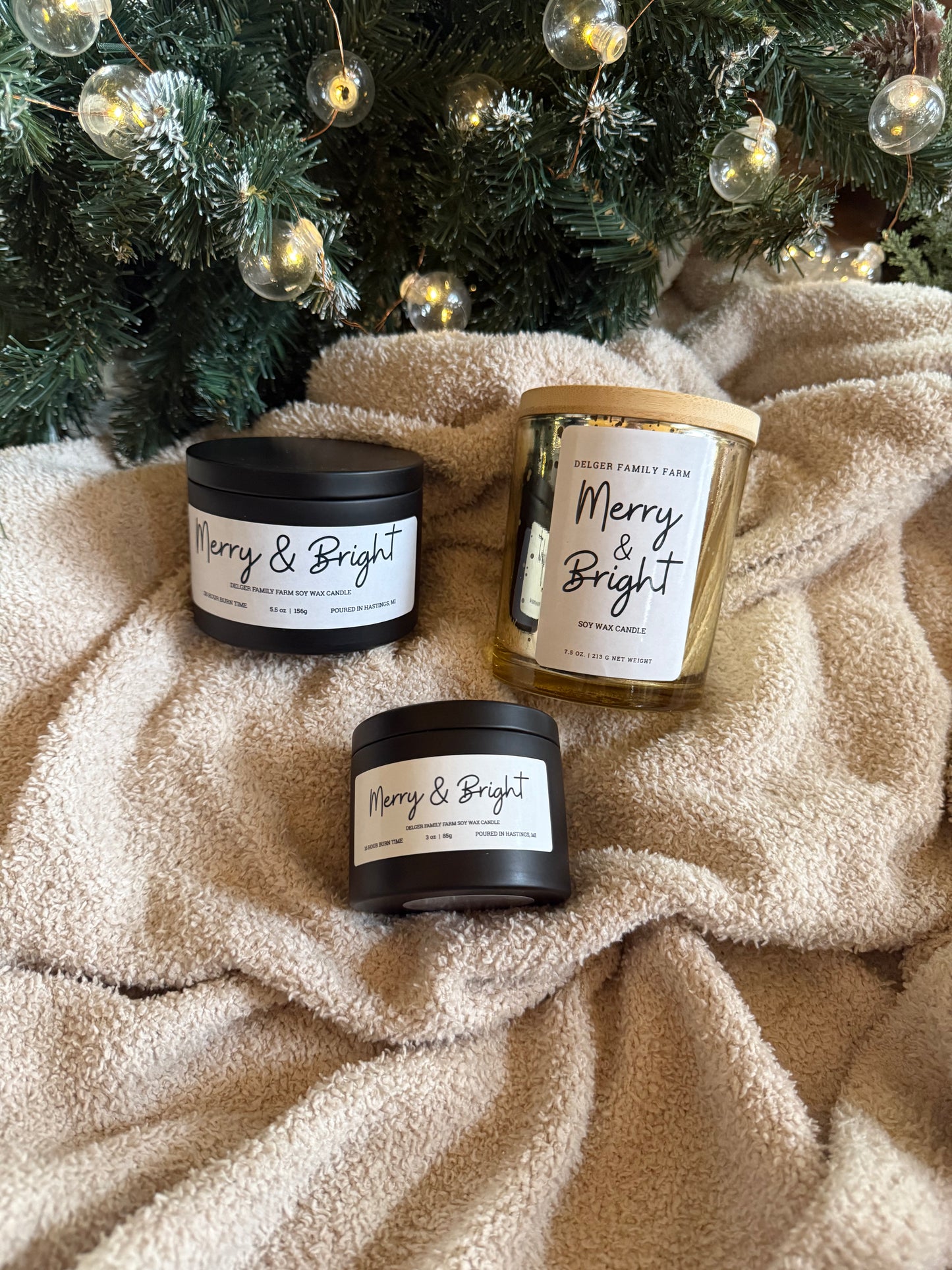 Three 'Merry & Bright' candles on a textured surface with a Christmas tree in the background.