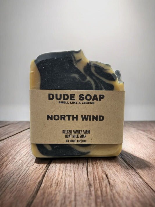 A rectangular bar of Dude Soap 'North Wind' goat milk soap, featuring a marbled black and yellow design with a kraft paper label, set on a rustic wooden surface against a neutral gray background.