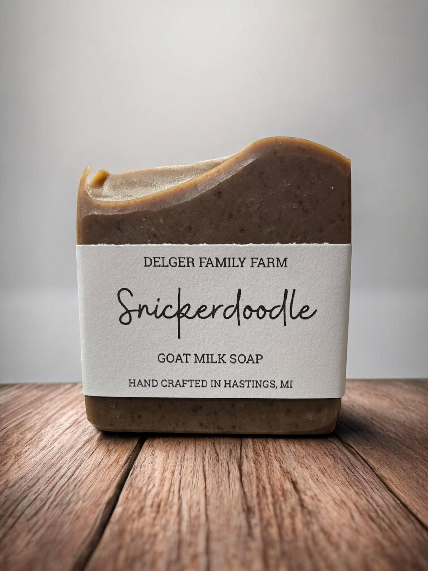 A rectangular bar of Delger Family Farm Snickerdoodle Goat Milk Soap, showing its textured brown top and bottom halves with a white paper label in the center, set on a rustic wooden surface against a neutral gray background.