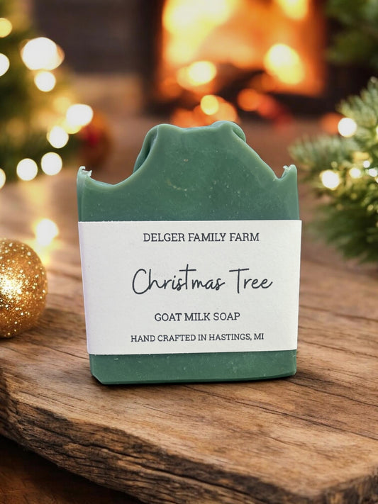 A bar of Delger Family Farm Christmas Tree goat milk soap in a dark green color, resting on a rustic wooden surface with a warm, cozy background featuring a blurred fireplace and holiday lights.