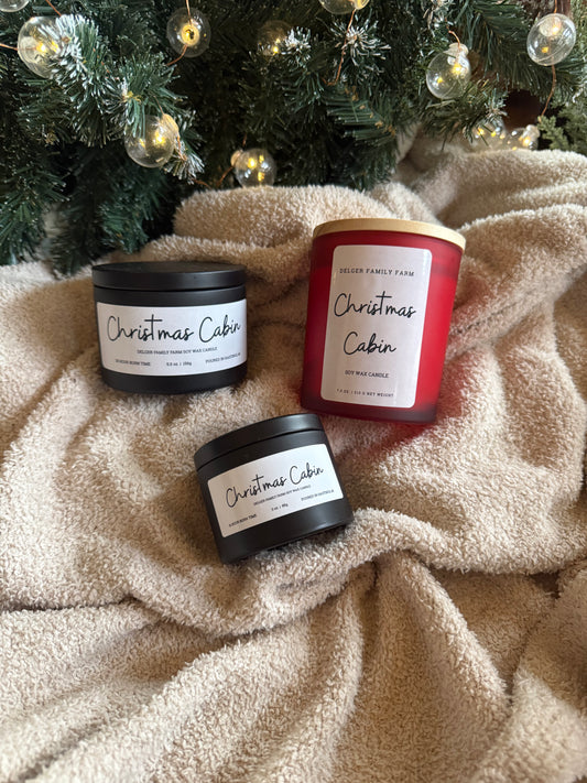Three candles labeled 'Christmas Cabin' on a soft blanket with a Christmas tree in the background.