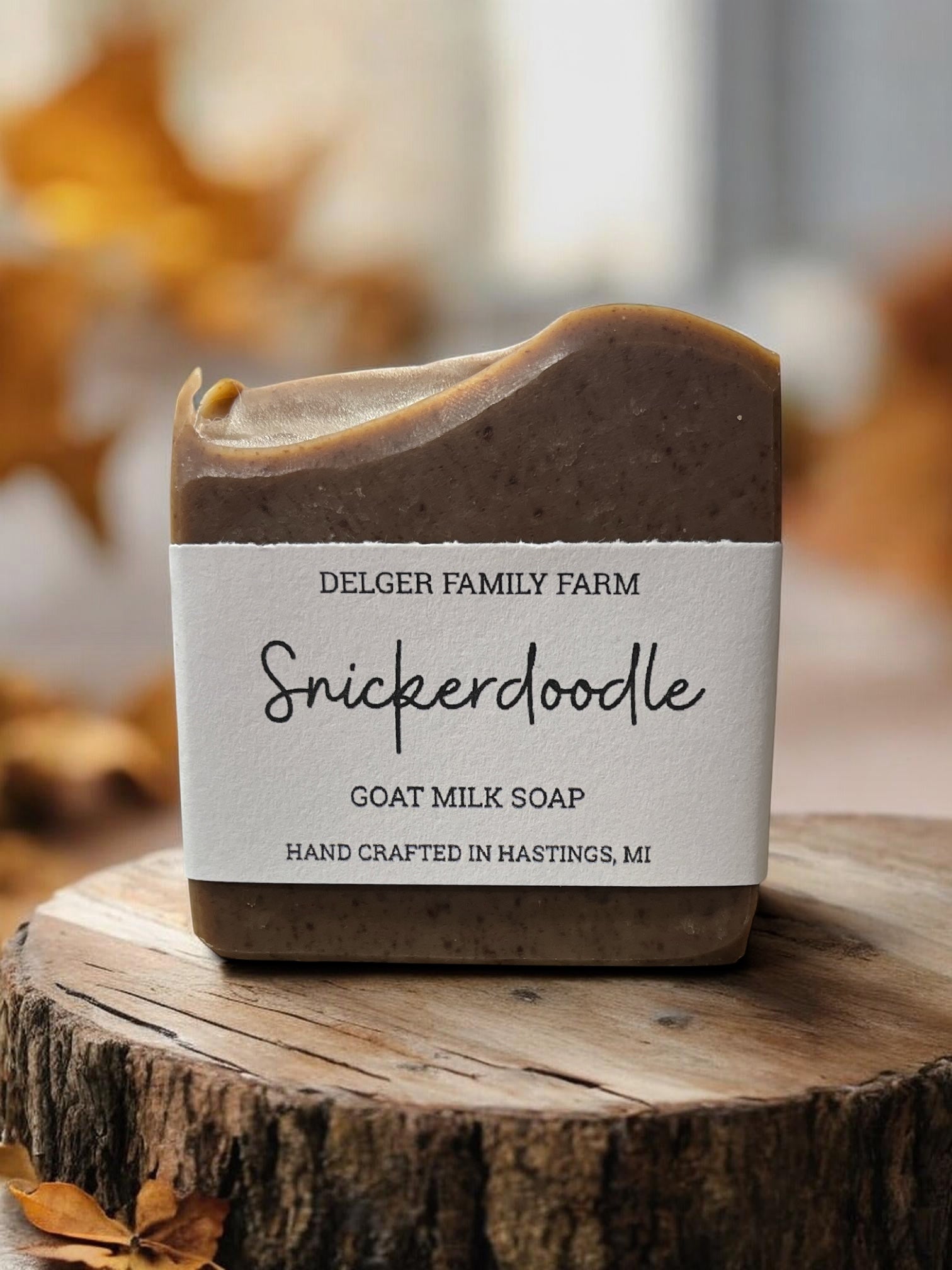 A bar of Delger Family Farm Snickerdoodle goat milk soap sits on a rustic wood slice. The soap has a warm brown top and bottom with a white paper label wrapped around the middle.