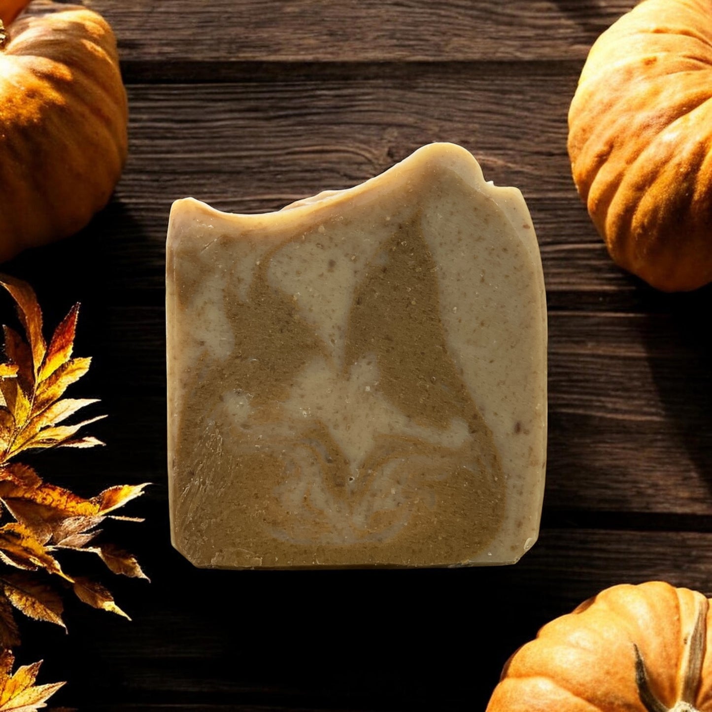 A rectangular bar of handmade soap with a marbled beige and light brown pattern, styled on a dark wooden background with decorative pumpkins and autumn leaves.