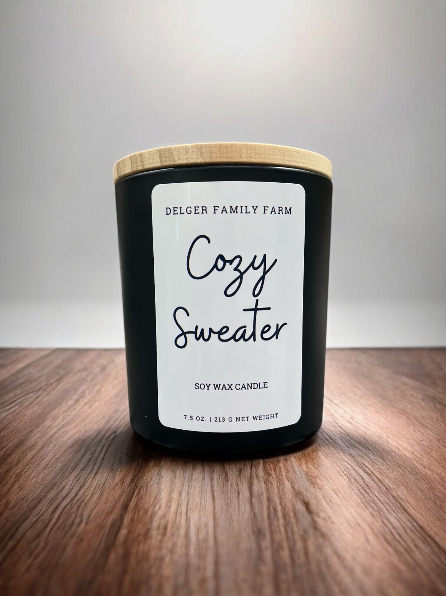 A black soy wax candle jar with a light wood lid and a white label reading 'Delger Family Farm Cozy Sweater,' placed on a wooden surface against a light gray background.