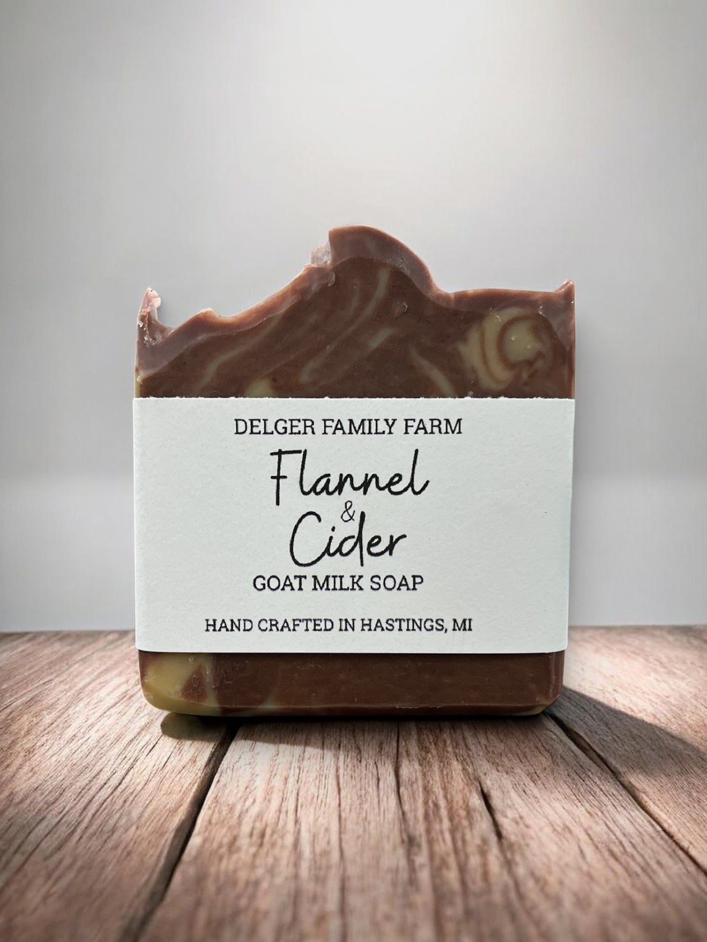 A bar of Delger Family Farm Flannel & Cider Goat Milk Soap, featuring a rustic hand-crafted top edge with visible swirls of brown and cream, set on a wooden surface against a neutral gray background.