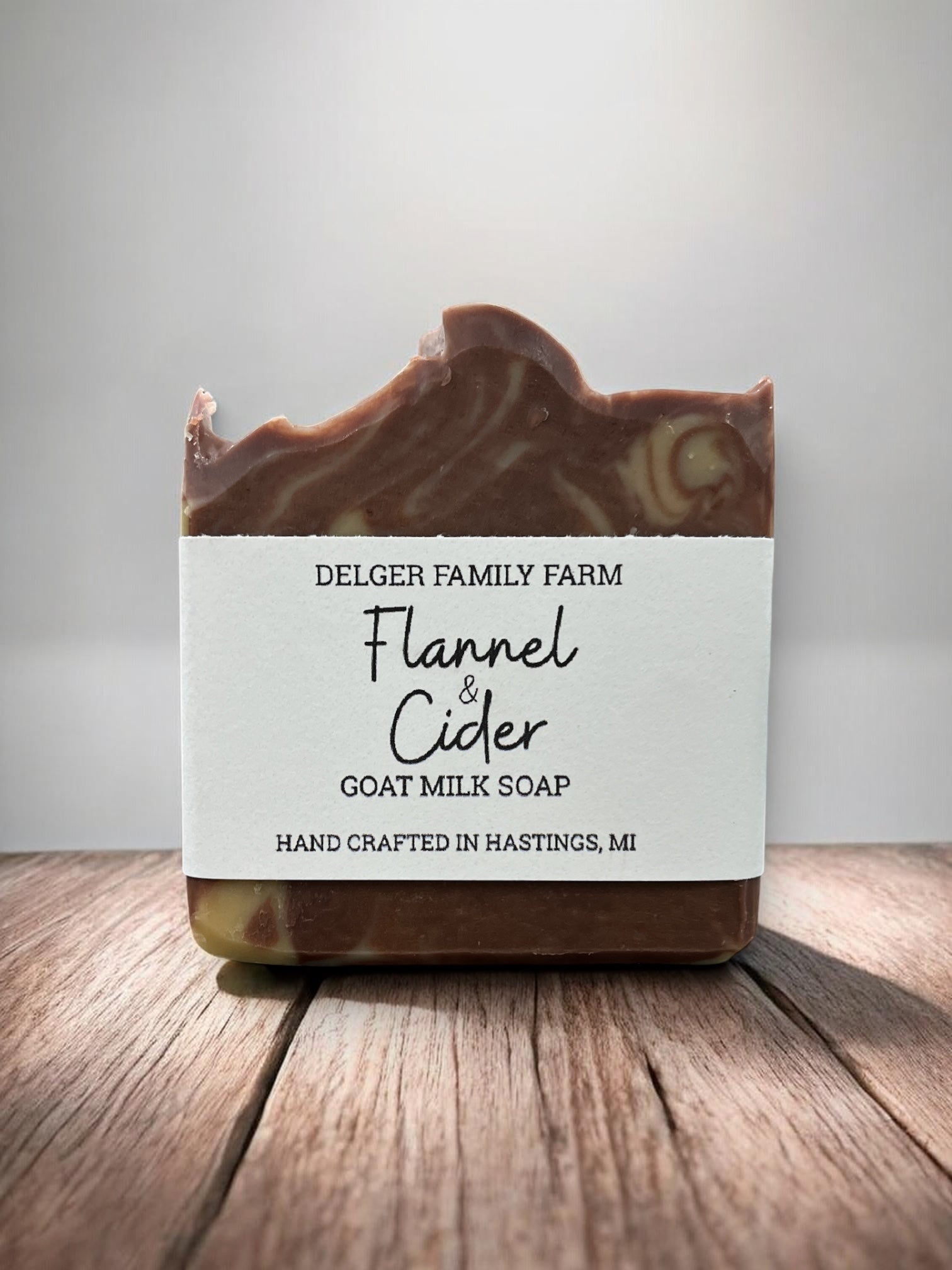 A bar of Delger Family Farm Flannel & Cider Goat Milk Soap, featuring a rustic hand-crafted top edge with visible swirls of brown and cream, set on a wooden surface against a neutral gray background.