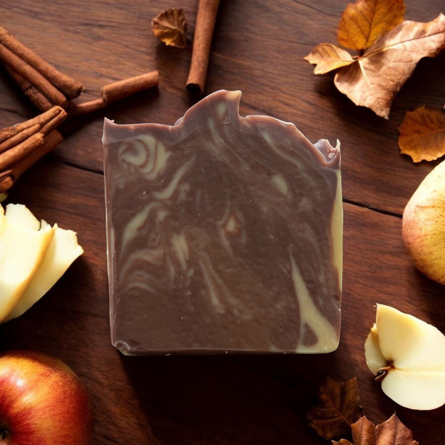 A rectangular bar of marbled soap with dark brown and cream swirls, styled on a dark wooden surface with cinnamon sticks, dried leaves, apple slices, and whole apples around it.