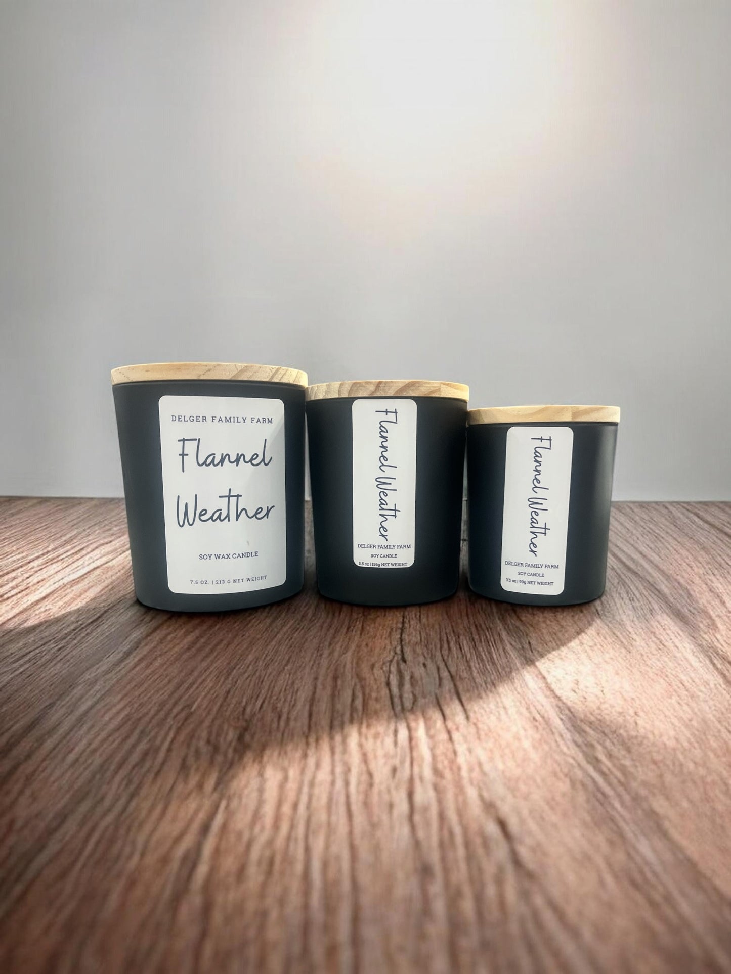 Three black soy wax candles with light wood lids are lined up on a wooden surface against a neutral gray background. The labels read 'Delger Family Farm' and 'Flannel Weather'.