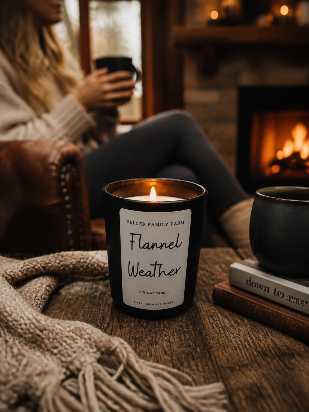 A lit black glass candle jar labeled 'Cedar Family Farm Flannel Weather' sits on a wooden table in a cozy living room setting with a person holding a mug in the background near a fireplace.
