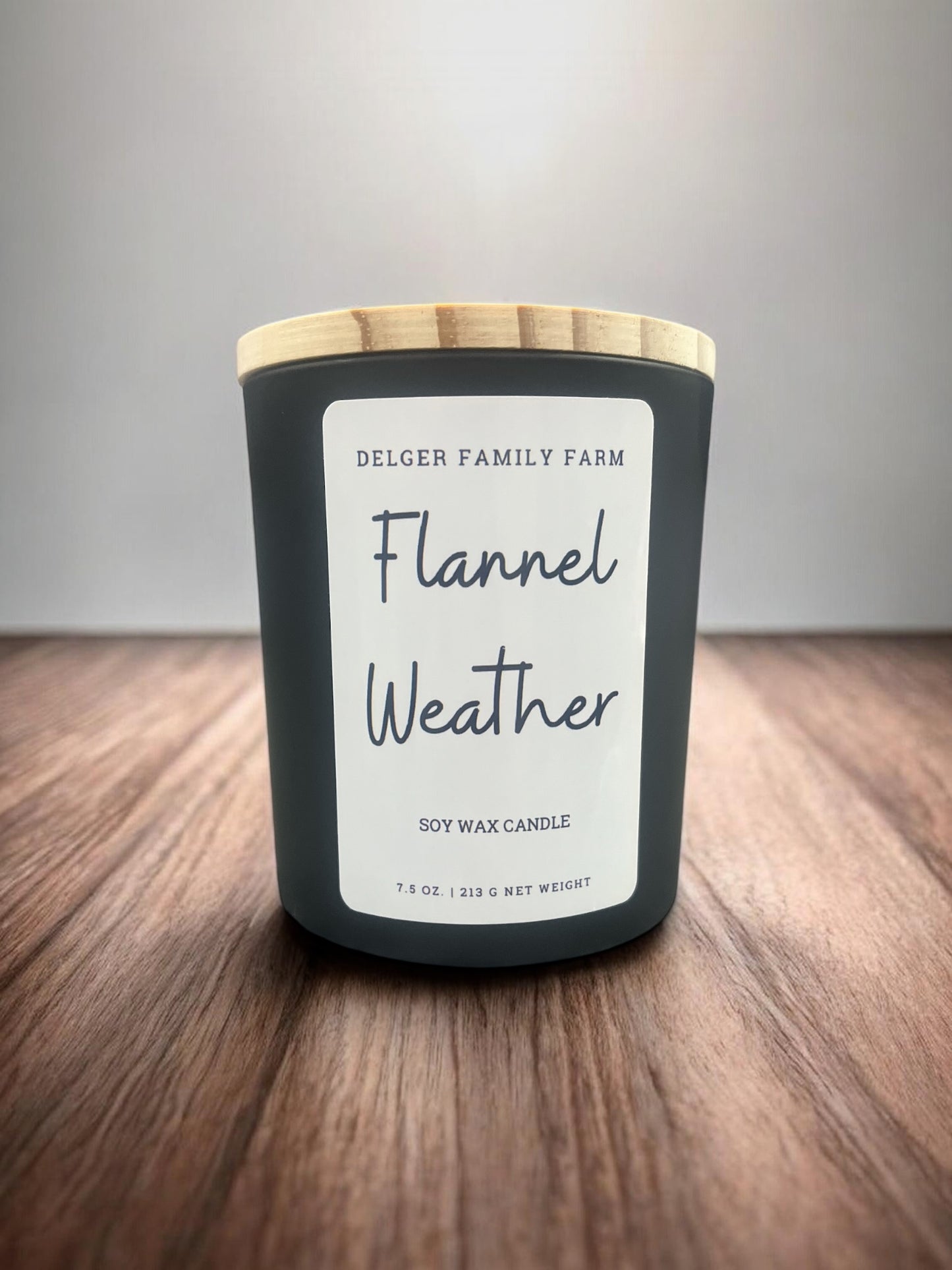 A soy wax candle in a matte dark gray jar with a light wood lid, labeled 'Delger Family Farm Flannel Weather,' placed on a wooden surface against a neutral gray background.