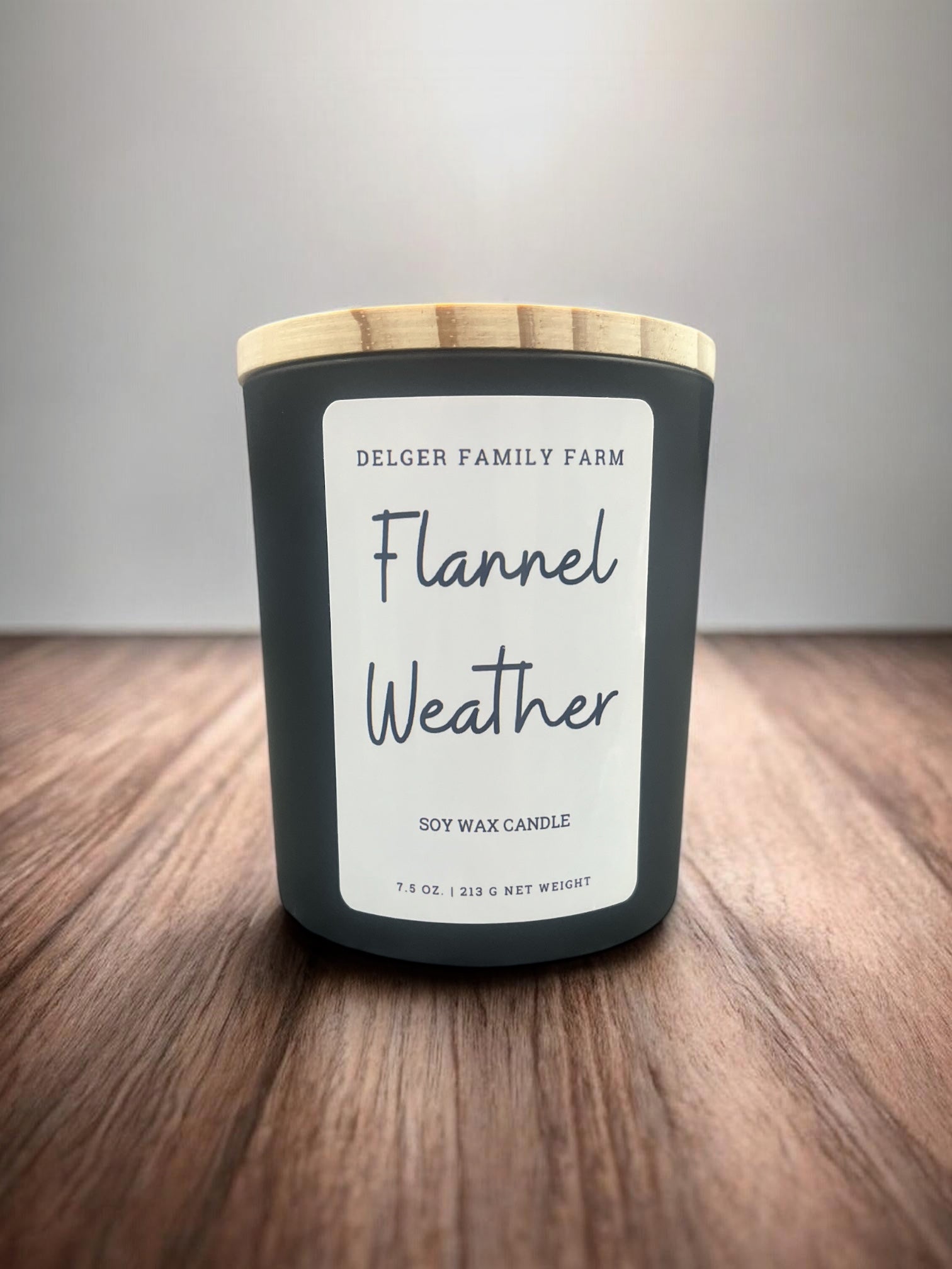 A soy wax candle in a matte dark gray jar with a light wood lid, labeled 'Delger Family Farm Flannel Weather,' placed on a wooden surface against a neutral gray background.