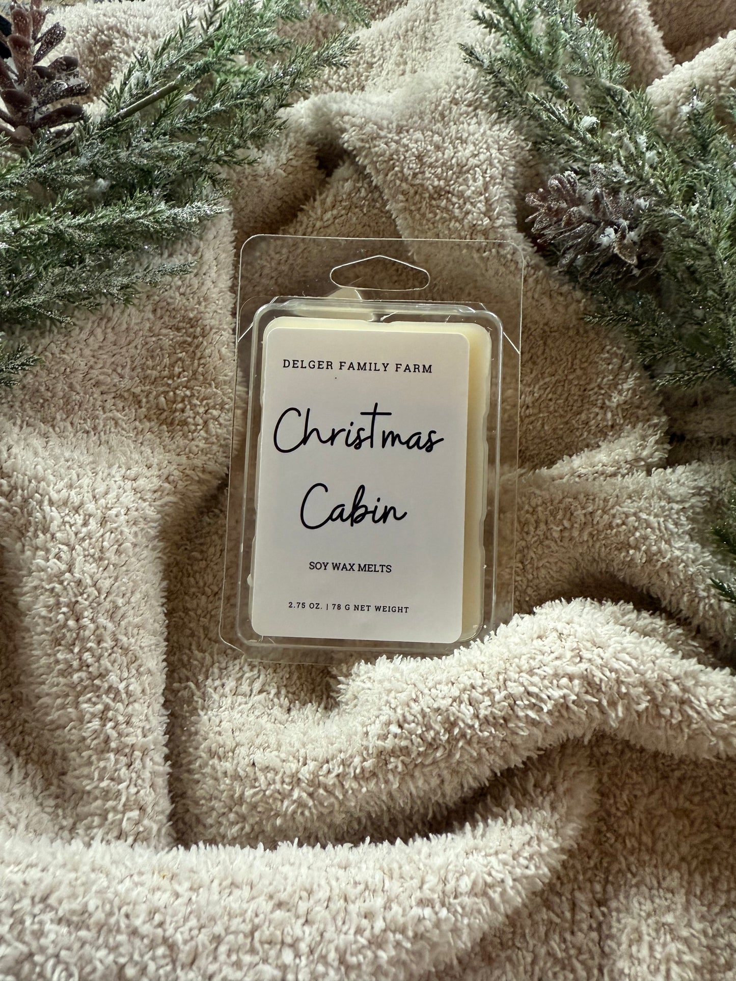 Packaged wax melts labeled 'Christmas Cabin' on a textured beige fabric background with greenery.