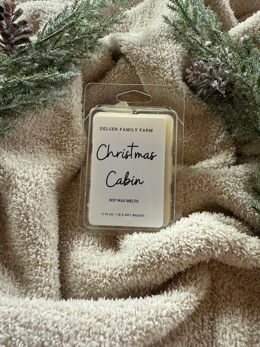 Packaged wax melts labeled 'Christmas Cabin' on a textured beige fabric background with greenery.