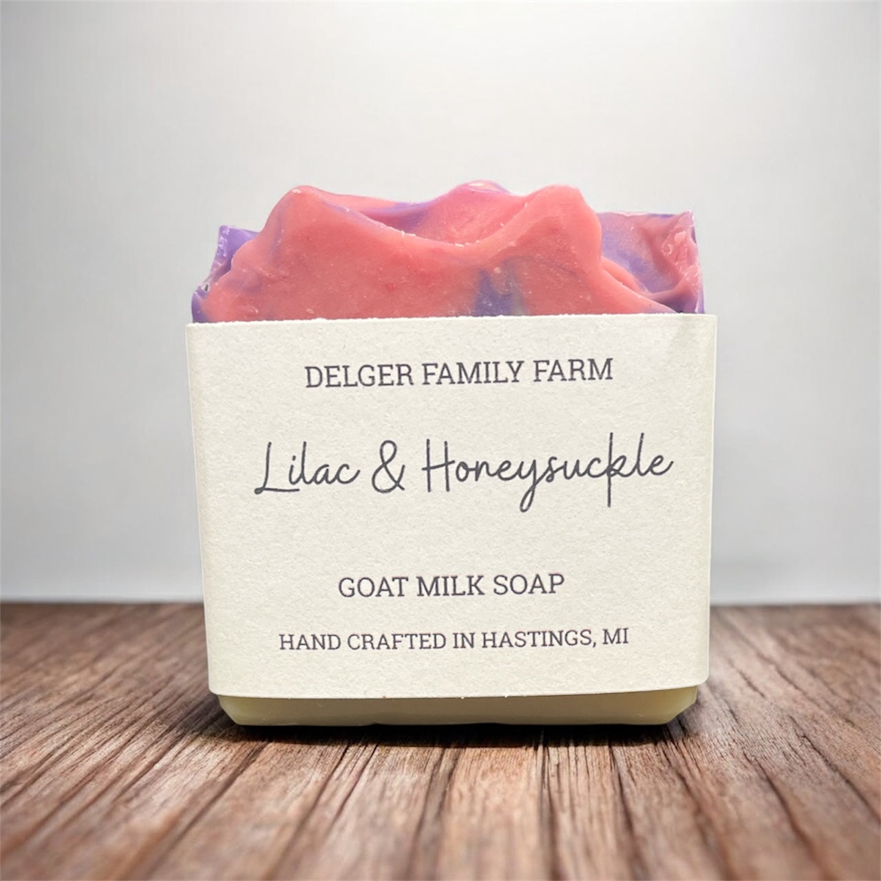 Bar of soap with a colorful design on top, labeled 'Delger Family Farm Lilac & Honeysuckle Goat Milk Soap' on a wooden surface.