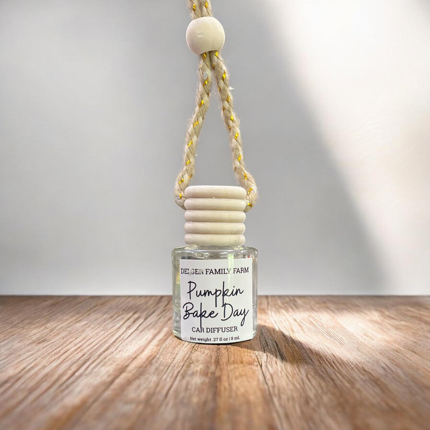 A glass car diffuser bottle with a white label reading 'Pumpkin Bake Day' and 'Golden Family Farm,' hanging from a braided rope with a round wooden bead against a light gray wall and wood floor.