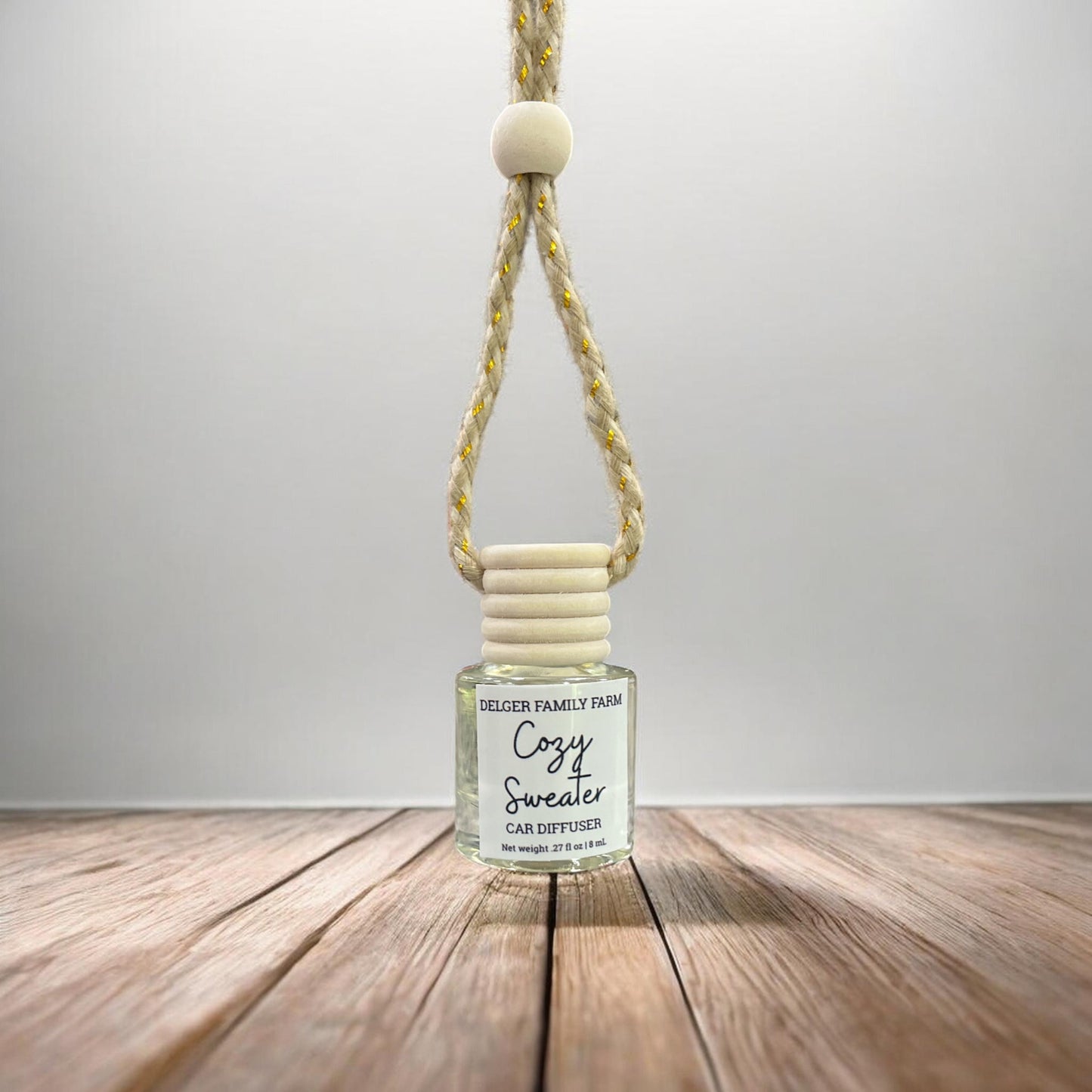 A Delger Family Farm 'Cozy Sweater' car diffuser with a clear glass bottle, cream-colored wooden cap, and a yellow-and-white braided hanging cord, displayed on a wooden surface against a neutral gray background.