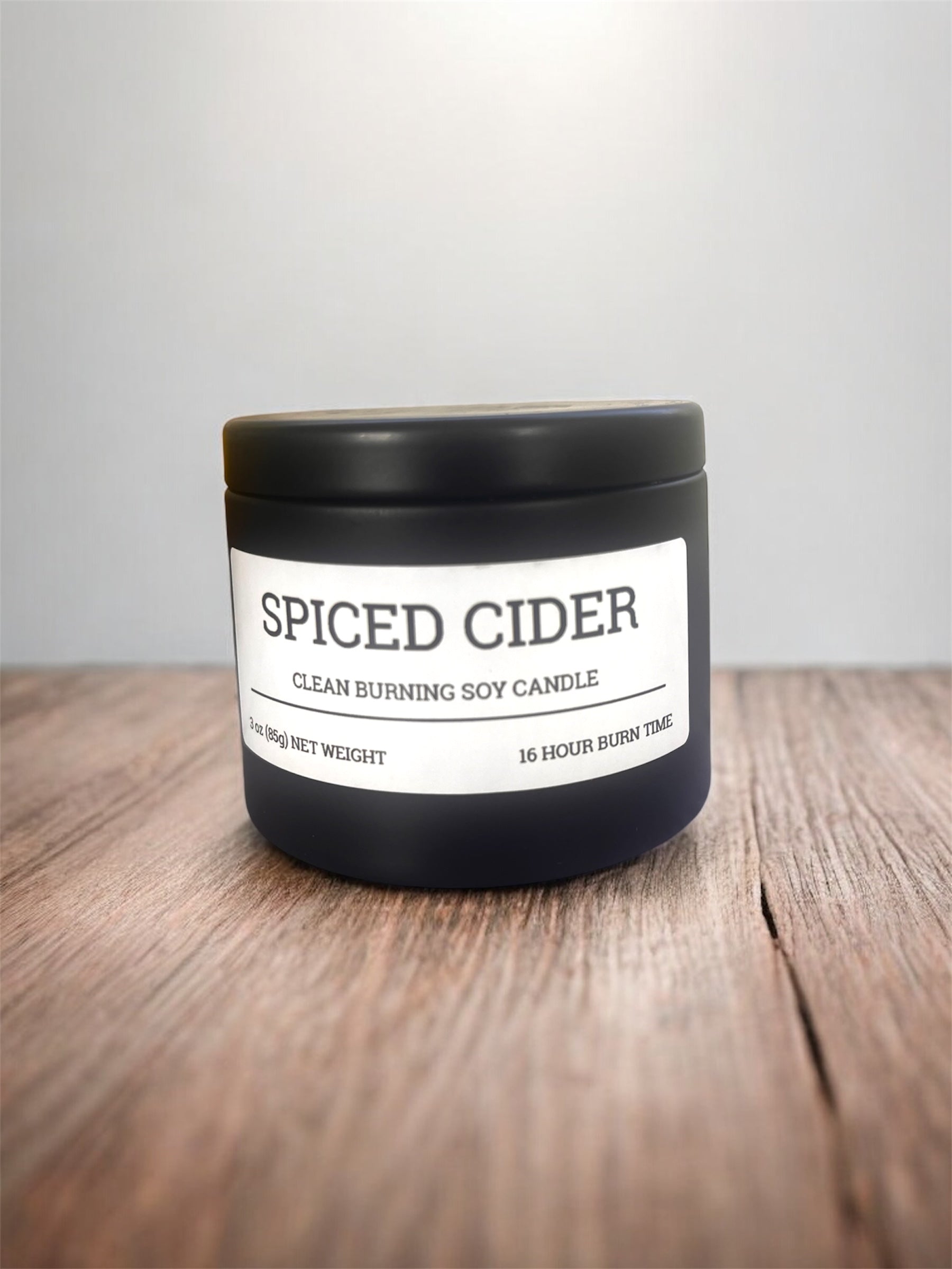 Spiced Cider candle in a black container on a wooden surface with a white background