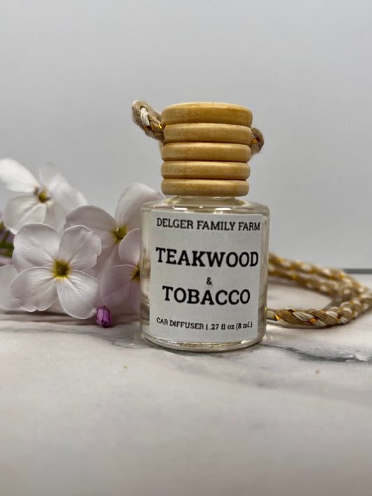 Small glass diffuser bottle labeled 'Teakwood & Tobacco' by Delger Family Farm on a marble surface with flowers in the background.