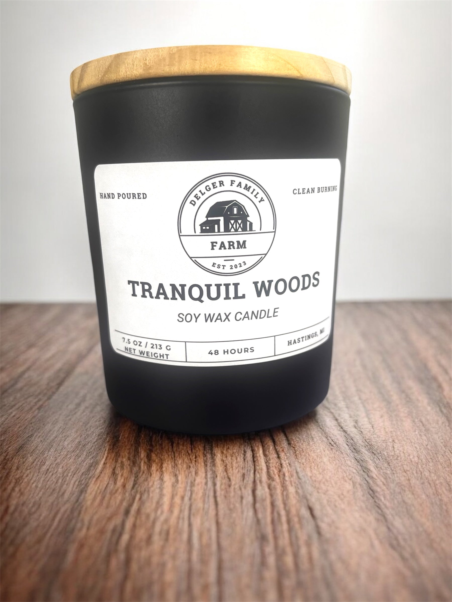 Black candle with a wooden lid labeled 'Tranquil Woods' on a wooden surface.